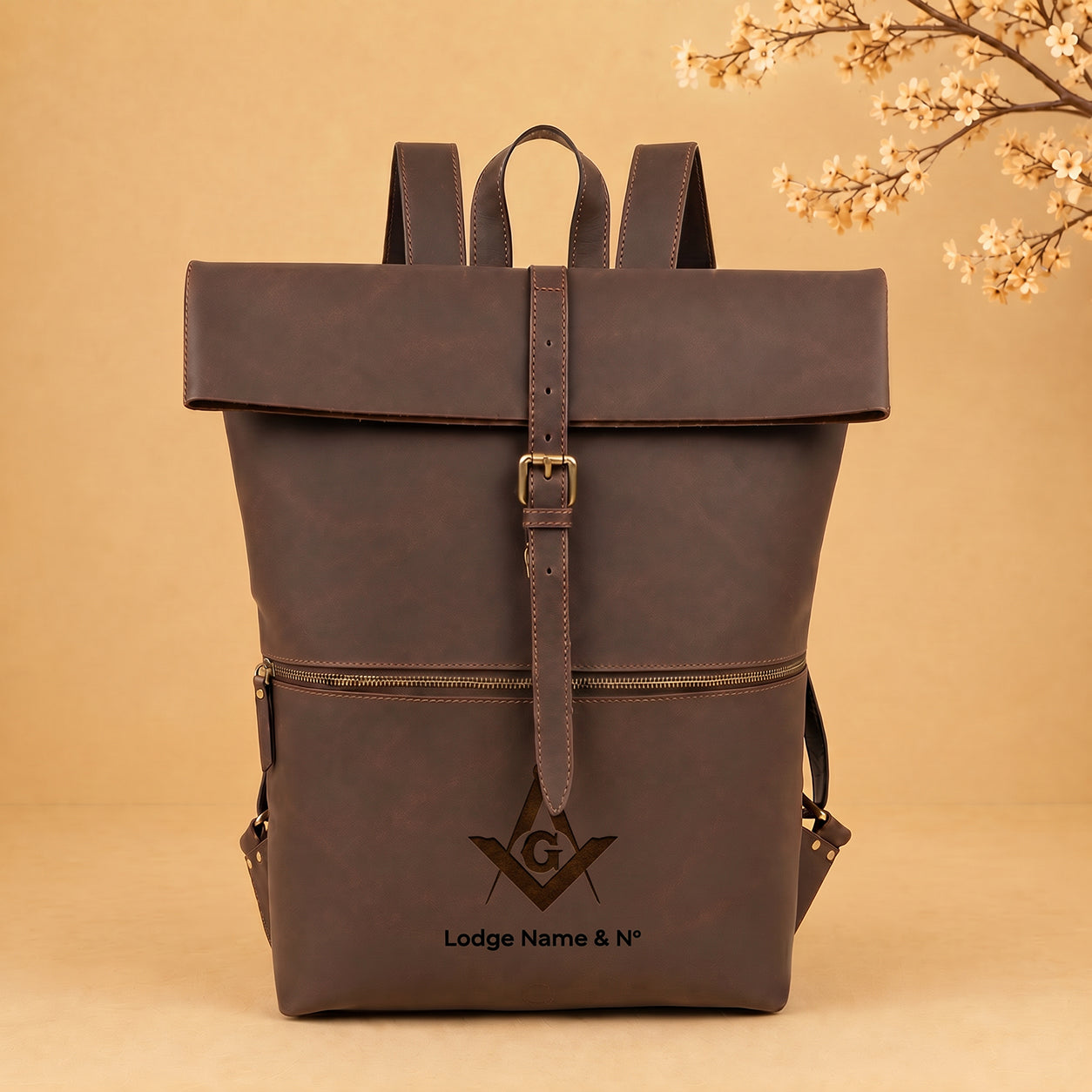 A dark brown leather backpack with a front flap, buckle strap, and a front zip pocket, featuring an embossed Masonic square and compass logo with the text 'Lodge Name & No' on the front, set against a warm beige background with decorative cherry blossoms in the top right corner.