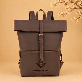A dark brown leather backpack with a front flap, buckle strap, and a front zip pocket, featuring an embossed Masonic square and compass logo with the text 'Lodge Name & No' on the front, set against a warm beige background with decorative cherry blossoms in the top right corner.